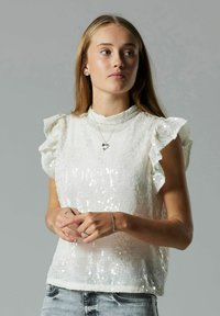 Valge sädelev top, millel on volangkätised ja kõrge kaelus, koos teksadega, näidates tekstuurset pinda ja peent sära.