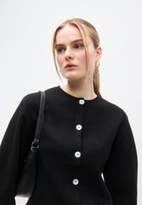 Μαύρο ζακέτα από υλικό με υφή με στρογγυλή λαιμόκοψη και μεγάλα λευκά κουμπιά. Μικρή μαύρη τσάντα κρατιέται στον έναν ώμο.
