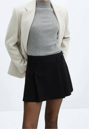 Vrouw die een beige blazer draagt, een grijze geribde coltrui, een zwarte geplooide rok en doorschijnende zwarte panty's, staand met handen in de blazerzakken.