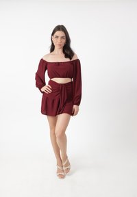 Mørkerød to-delt sæt; off-the-shoulder top med lange ærmer og cropped design, parret med et knyttet mini nederdel. Neutrale strophæle.