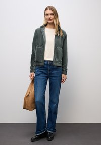 Green velour zip-up jacket with two front pockets, paired with light cream top and blue flared jeans. Brown suede tote bag, black ankle boots.