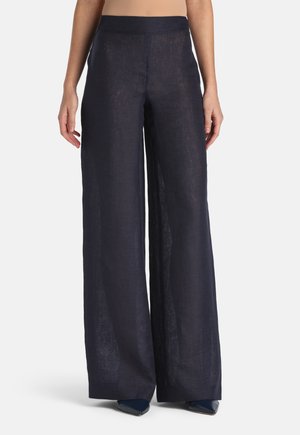 Kobieta ubrana w granatowe lniane spodnie o wysokim stanie i szerokich nogawkach, w połączeniu z granatowymi spiczastymi butami, stojąca na tle jednolitego jasnego tła.