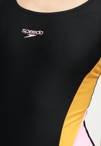 Μαύρο μαγιό Speedo με κίτρινες και ροζ πλευρικές επιφάνειες, που δείχνει το πάνω μέρος του σώματος και το λογότυπο στο στήθος.