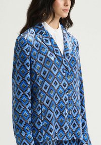 Haut de pyjama bleu à motifs avec des formes losanges en bleu clair, vert et noir. Fini satin avec col cranté et fermeture à boutons.