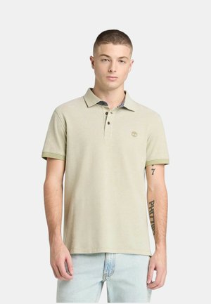 Giovane uomo in piedi che indossa una polo beige a maniche corte con bottoni e jeans azzurri chiari su uno sfondo uniforme.