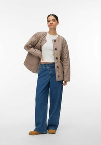 Bež prešit jakna s črnimi gumbi, kombinirana s širokimi modrimi kavbojkami in rjavimi čevlji, postavljena na preprosto belo ozadje.