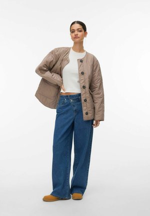 Veste matelassée beige avec des boutons noirs, associée à un jean bleu à jambes larges et des chaussures marron clair, sur un fond blanc uni.