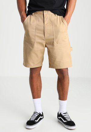 Hombre vestido con pantalones cortos beige hasta la rodilla, camiseta negra, calcetines blancos y zapatillas negras con cordones blancos, de pie con las manos en los bolsillos.