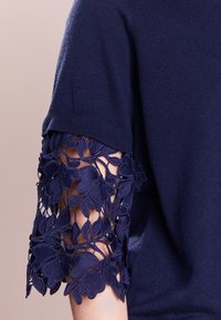 Suéter de punto azul marino con acentos de encaje floral en las mangas cortas. El encaje tiene un diseño texturizado y en capas.
