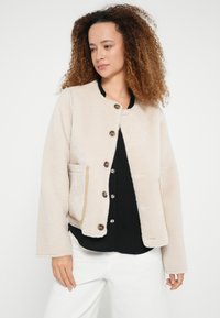 Beżowa bluza polarowa z okrągłym dekoltem, guzikami z przodu i bocznymi kieszeniami. Charakteryzuje się ozdobnymi przeszyciami i miękką fakturą.