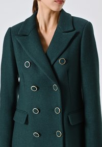 Teal dubbelzijdige jas gemaakt van gestructureerde stof, met gouden ronde knopen, een gesneden kraag en zijzakken.
