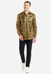 Langärmliges Hemd mit gold- und schwarzem Muster im Schlangenlederdesign, kombiniert mit schwarzen Hosen und weißen Sneakers. Glattes, glänzendes Material.
