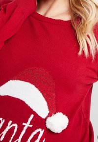 Red sweater featuring a white Santa hat graphic with a fluffy white pom-pom. Material appears soft with a textured finish.