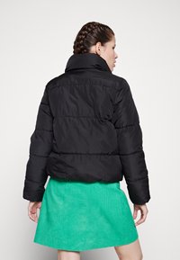 Svart pufferjakke med høy krage, rutete design og avrundet kant, kombinert med et grønt corduroy-skjørt.