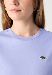 Šviesiai violetinė marškinėliai su apvalia iškirpte ir mažuoju išsiuvinėtu žaliu krokodilo logotipu kairėje krūtinės pusėje. Minkšta medvilnė.