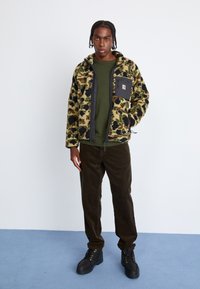 Veste en polaire avec un motif camouflage et un panneau avant noir, associée à un pantalon en velours côtelé sombre et des bottes noires à lacets.