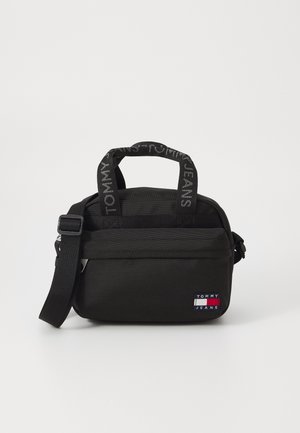 Schwarze Stofftasche mit kurzen Henkeln, verstellbarem Riemen, Reißverschluss und einem kleinen Logopatch in Rot, Weiß und Blau.