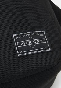 Fekete szövet táska texturált felülettel, egy téglalap alakú címkével, fehér varrással, amelyen a "PIER ONE" és a "PRÉMIUM MINŐSÉGŰ TERMÉKEK" felirat olvasható.