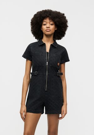 Femme aux cheveux bouclés vêtue d'une combinaison courte noire à manches courtes avec fermeture éclair à l'avant, poches poitrine et bretelles latérales ajustables, posant devant un fond blanc.