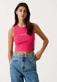 Aansluitend geribbelde tanktop in felroze, met een ronde hals en witte "RED BULL RACING" logo. Gecombineerd met high-waisted denim jeans.