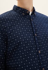Navyblaues Hemd mit Knöpfen und weißen Mustern, mit einem klassischen Kragen, braunen Knöpfen und einer glatten Stofftextur.