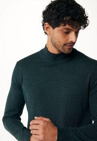 Tmavě zelený pletený svetr s vysokým límcem, dlouhými rukávy a ribbovanými manžetami. Tkanina vypadá jemně a má hladký povrch.