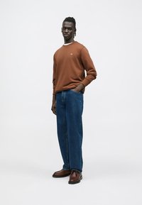 Sweat-shirt marron avec un col blanc, associé à un jean bleu ample et des bottines marron. Le mannequin pose avec une main dans la poche.