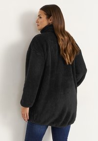 Schwarze, oversized Fleecejacke mit hohem Kragen, versehen mit einer weichen, strukturierten Oberfläche und überschnittenen Schultern, getragen mit blauen Jeans.