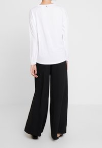 Vitt långärmad skjorta med rund halsringning och en liten dekorativ knapp på baksidan, kombinerad med svarta vida byxor.