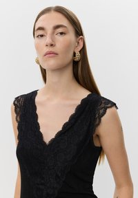 Svart ärmlös top med djup V-ringning, försedd med texturerad spetskant vid axlar och halsringning. Modellen bär guldörhängen i form av ringar.