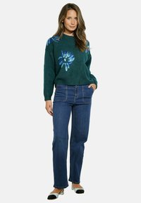 Pullover verde a maglia con design floreale in paillettes blu, abbinato a jeans a gamba dritta blu e scarpe bicolore nere e azzurre.
