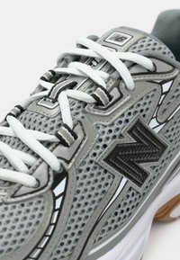 Sneaker grigio New Balance con lacci bianchi, tomaia in rete, logo "N" nero e asole per lacci con motivo su sfondo bianco.