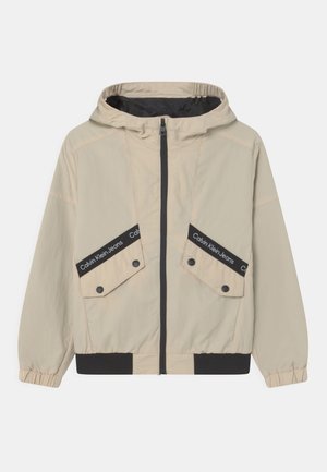 Giacca leggera beige con cappuccio, cerniera nera e polsini elastici. Presenta due ampie tasche frontali con cinturini neri brandizzati.