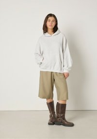 Sudadera con capucha gris y bolsillo canguro, combinada con pantalones cortos oliva hasta la rodilla y botas de cuero marrón hasta la rodilla.