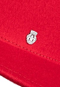 Red felt cap with stitched edge; features a silver crest badge with a detailed design. Smooth texture with a vibrant color.
