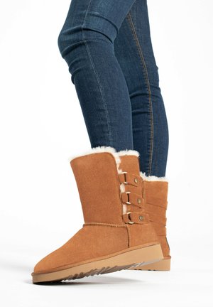 Tan suede ankle boots with white faux fur lining, featuring three decorative buttons and a flat rubber sole. Styled with dark skinny jeans.