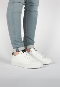 Sneaker bianchi in pelle con una texture liscia, dotate di lacci bianchi, un accento nero sul tallone e una suola in gomma, abbinate a jeans blu chiaro.
