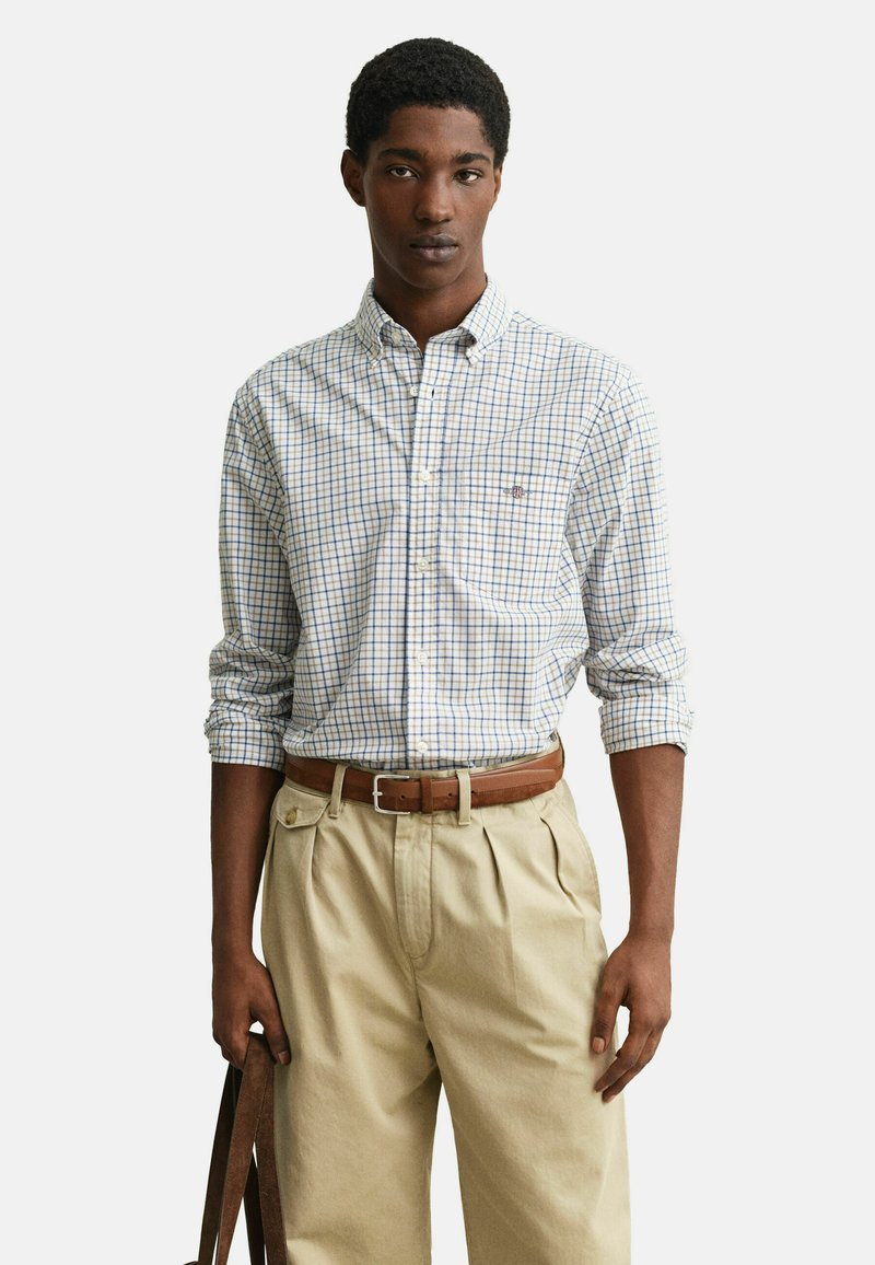 Jeune homme portant une chemise à carreaux blanche et bleue à boutons, manches retroussées, pantalon beige plissé, ceinture marron, tenant les anses d'un sac marron.