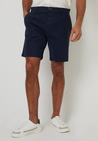 Shorts en coton bleu marine avec poches latérales, fermeture à bouton et coupe droite. Portés avec des baskets blanches à accent gris.