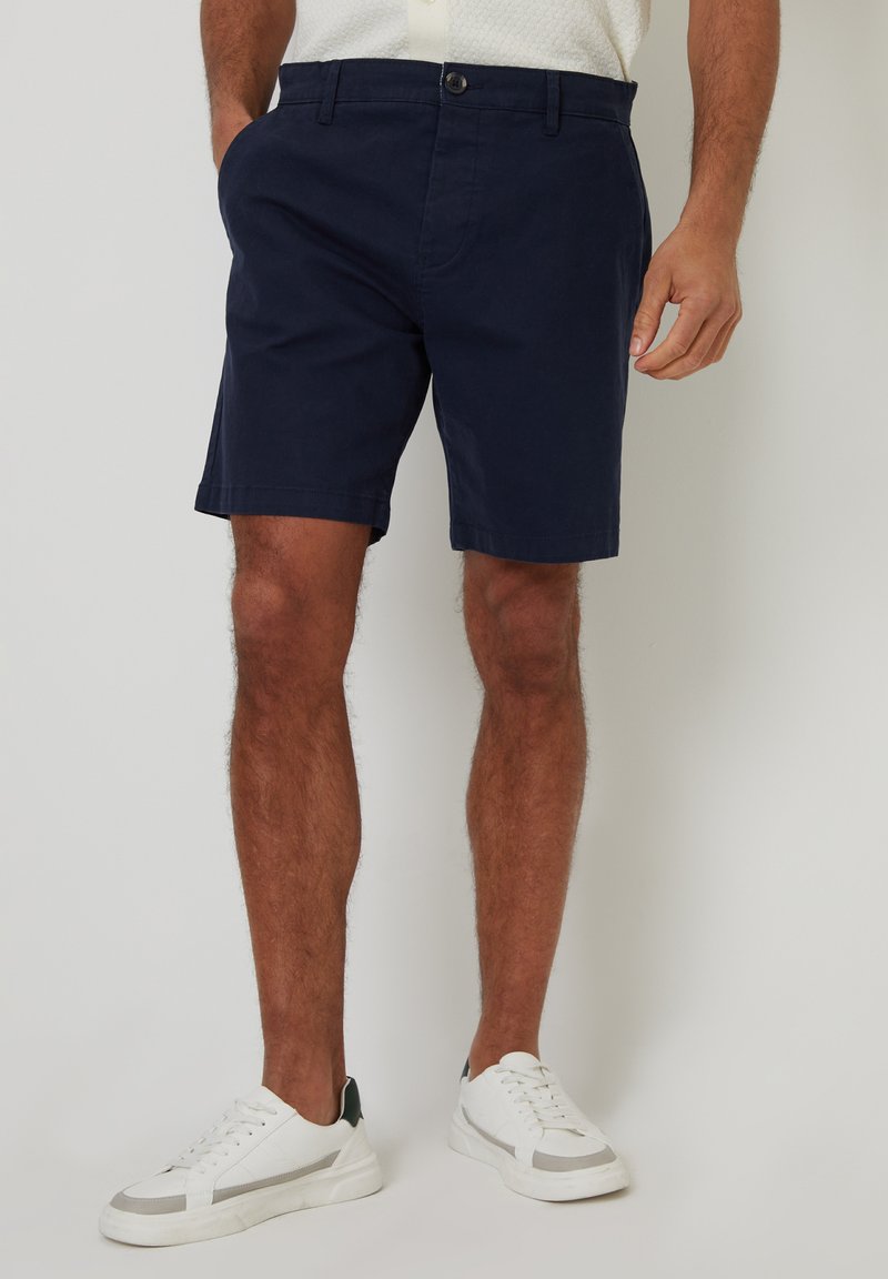 Shorts en coton bleu marine avec poches latérales, fermeture à bouton et coupe droite. Portés avec des baskets blanches à accent gris.