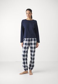 Marinblå långärmad topp i kombination med rutiga pyjamasbyxor i blått och vitt, med elastiska mudd i ärmarna och en avslappnad passform. Modellen står barfota.