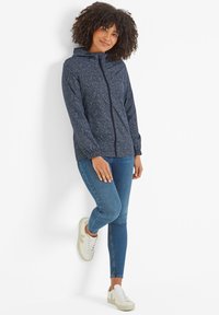 Navy jacket with a hood and intricate white pattern, featuring a front zipper and elastic cuffs. Paired with blue jeans and white sneakers.