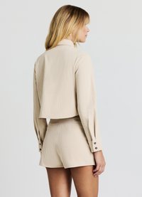 Giacca corta beige con texture a coste e maniche lunghe, abbinata a pantaloncini coordinati con vita alta e dettagli con bottoni.