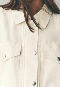 Beige denimjakke med krave, der har sølvfarvede trykknapper og to brystlommer. Stoffet har en glat, let struktureret overflade.