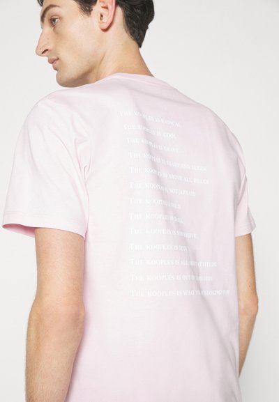 Kortermet rosa t-skjorte laget av myk stoff, med gjentatt hvit tekst på ryggen med diverse bekreftelser fra "The Kooples."