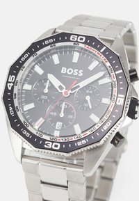 Reloj de acero inoxidable plateado con un dial negro, sub-diales de cronógrafo, marcadores de hora luminosos y un bisel taquimétrico con acentos rojos.