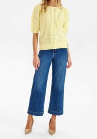 Gul stickad tröja med puffärmar, kombinerad med vida blå denimjeans och nude-hälar, vilket visar en avslappnad outfit.