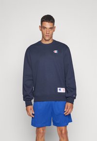 Sudadera navy con cuello redondo, puños y dobladillo acanalados, que presenta un pequeño logo en el pecho y una etiqueta en la parte inferior izquierda. Combinada con pantalones cortos azules.