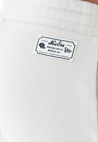 Weißer Stoff mit strukturierter Oberfläche und gestepptem Saum, versehen mit einem rechteckigen New Era-Label in Schwarz und Weiß mit Markendetails.