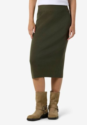Femme portant une jupe midi côtelée vert olive et des bottines en daim beige avec des sangles, debout devant un fond blanc.